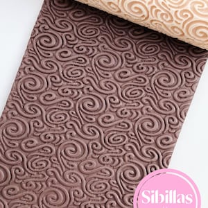 Könnte beinhalten: Ein beiger Nudelholz mit einem Wirbelmuster liegt auf einer braunen Oberfläche mit einem passenden geprägten Design. Das Nudelholz besteht aus einem hellfarbigen Material, und die Oberfläche ist ein dunklerer Braunton. Der Text "Sibillas cutters" ist enthalten.