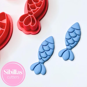 May include: Red and blue mermaid tail cookie cutters and two blue mermaid tail shapes on a white surface. The cutters are plastic and the shapes are made of a blue material. The image includes the text "Sibillas cutters" in a pink circle.