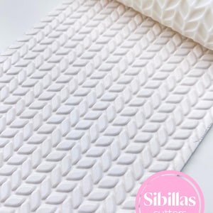 Op de afbeelding: Een witte, getextureerde deegroller met een herhalend ruitpatroon. De deegroller is gedeeltelijk opgerold, waardoor het patroon zichtbaar is. De afbeelding bevat een roze cirkel met de tekst "Sibillas cutters".