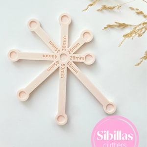 May include: A set of light pink cookie cutters in various sizes, arranged in a starburst pattern. Each cutter has a circular handle and is labeled with a measurement in millimeters, ranging from 20mm to 55mm. The Sibillas Cutters logo is visible.
