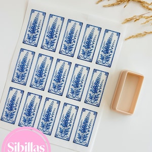May include: A sheet of rectangular paper featuring fifteen blue and white floral illustrations of delphinium flowers in a tile-like pattern, accompanied by a single rectangular beige plastic cookie cutter positioned to the right of the paper.