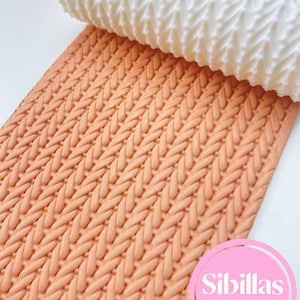 Puede incluir: Un rodillo blanco texturizado se muestra parcialmente enrollado sobre una lámina de fondant color melocotón, creando un patrón de punto. La imagen presenta una vista de cerca de la superficie texturizada, ideal para decorar pasteles. El logotipo de Sibillas Cutters es visible.
