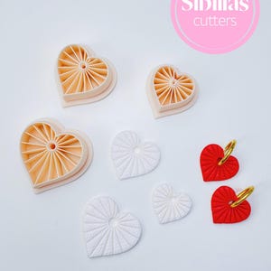 Peut inclure: Diverses découpoirs et décorations en forme de cœur. Les découpoirs sont de couleur pêche clair, tandis que les décorations sont blanches et rouges. L'image comprend le texte "Sibillas cutters" dans un cercle rose.