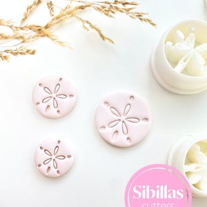May include: Pink sand dollar-shaped cookie cutters and white plastic molds are displayed on a white surface. The cutters have a detailed design. The image also includes a logo that says "Sibillas cutters".