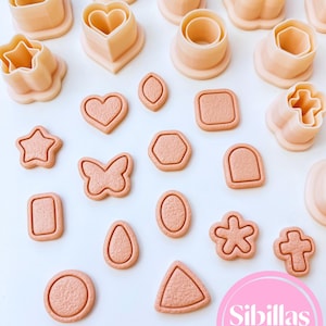 Peut inclure: Une collection d'emporte-pièces de couleur pêche clair de différentes formes, notamment des étoiles, des cœurs, des papillons et des formes géométriques. Les emporte-pièces mesurent 1,5 cm et sont disposés sur une surface blanche. Le texte "Sibillas cutters" est inclus.