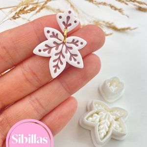 Può includere: Un orecchino bianco a forma di fiore con intagli complessi e un anellino dorato, accompagnato da due stampini per biscotti in plastica bianca con lo stesso motivo floreale. Il testo Sibillas cutters è visibile in un cerchio rosa.