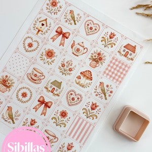 Puede incluir: Una hoja rectangular con una cuadrícula de pequeñas ilustraciones cuadradas en tonos coral y melocotón, que incluyen pájaros, flores, teteras, casas, corazones y lazos. Un pequeño recipiente cuadrado de plástico de color claro se encuentra junto a la hoja.
