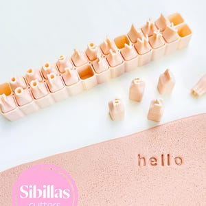 Puede incluir: Un juego de cortadores de alfabeto rosa claro con letras individuales, dispuestos en una bandeja. La palabra "hello" está impresa en una superficie rosa texturizada. La imagen también incluye un círculo rosa con el texto "Sibillas cutters".