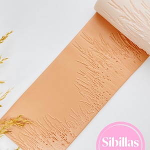 May include: A light peach-colored rolling pin with a wheat stalk pattern is partially rolled out on a matching peach-colored surface. The rolling pin is on a white background. The image includes the text "Sibillas cutters" in a pink circle.