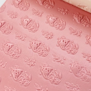 May include: A pink rolling pin with a repeating pattern of embossed strawberries and floral accents. The rolling pin is partially rolled out on a matching pink surface, revealing the textured design. The rolling pin is for baking.