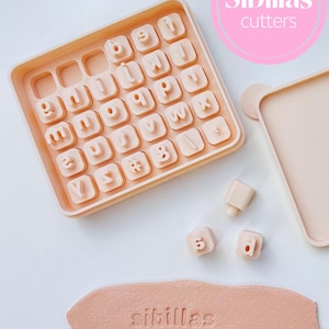 Op de afbeelding: Een licht perzik kleurige alfabet koekjessnijder set met individuele letter stempels. De set bevat een bakje met lettervormen, losse stempels en een stuk deeg met de tekst "sibillas cutters".