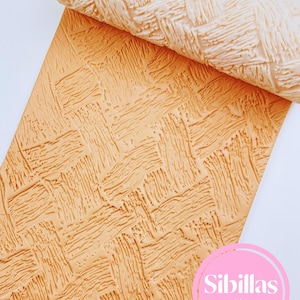 Può includere: Un mattarello beige con una trama intrecciata è parzialmente arrotolato su un foglio abbinato. Il foglio e il mattarello hanno un motivo a trama di cesto. L'immagine include un cerchio rosa con il testo "Sibillas cutters".