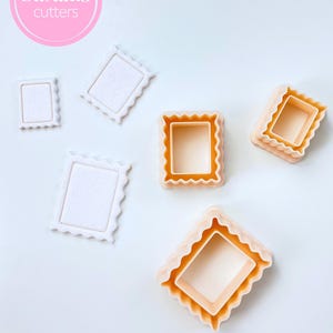 Pode incluir: Uma coleção de cortadores de biscoitos em forma de moldura nas cores branca e pêssego. Os cortadores têm um design de borda ondulada e são dispostos em uma superfície branca. A imagem inclui um círculo rosa com o texto "Sibillas cutters".