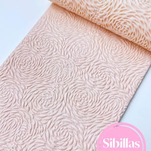 Könnte beinhalten: Rolle aus hellrosa Material mit einem sich wiederholenden Rosenmuster. Das Material ist mit erhabenen Wirbeln strukturiert, wodurch ein dreidimensionaler Effekt entsteht. Der Text "Sibillas cutters" ist in einem rosa Kreis sichtbar.