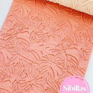 May include: A coral-colored rolling pin and a sheet of textured material, both featuring a repeating leaf pattern. The rolling pin is a light beige color. The sheet of material has a raised leaf design. The text "Sibillas cutters" is in a pink circle.