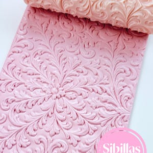 Könnte beinhalten: Ein hellrosa zylindrisches Nudelholz mit erhabenem floralem Rankenmuster, positioniert über einer passenden rosa flachen Materialplatte, die mit demselben komplexen, sich wiederholenden botanischen Design geprägt ist. Ein rosa kreisförmiges Logo zeigt Sibillas cutters.
