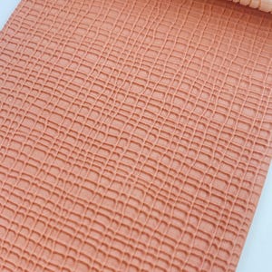 May include: Close-up of a roll of textured, coral-colored faux leather fabric. The material has a grid-like pattern, resembling a crocodile or alligator skin. The rolled portion at the top is a lighter, cream color.