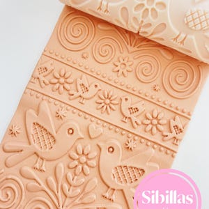Könnte beinhalten: Eine beige Teigrolle mit einem detaillierten Vogel- und Blumenmuster. Das Muster ist auf einer passenden beigen Oberfläche geprägt, die Vögel, Blumen und wirbelnde Designs zeigt. Das Bild enthält den Text "Sibillas cutters" in einem rosa Kreis.