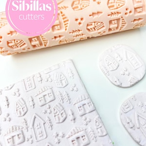 May include: A light pink rolling pin with a village pattern and the text "Sibillas cutters" in a pink circle. Also shown are light pink cookie cutouts and a square of light pink dough with the same village pattern.