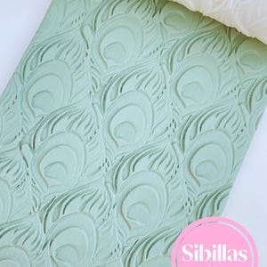 Puede incluir: Rodillo de amasar verde claro con un diseño en relieve de plumas de pavo real. El rodillo está parcialmente desenrollado, revelando un diseño blanco en relieve. El texto "Sibillas cutters" está en un círculo rosa. Para repostería.