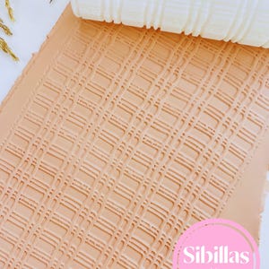 Pode incluir: Um rolo branco texturizado é pressionado em uma folha de argila bege, criando um padrão de grade. A folha de argila é impressa com um design retangular repetitivo. A imagem inclui o texto "Sibillas cutters" em um círculo rosa.