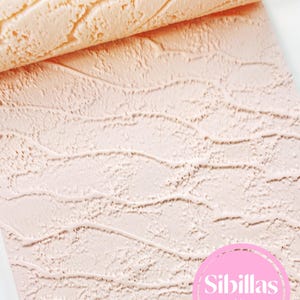 Pode incluir: Uma folha texturizada de cor pêssego claro com um padrão repetitivo de linhas orgânicas onduladas em relevo que lembram dunas de areia ou água corrente. Um logótipo circular rosa com o texto branco Sibillas cutters está no canto inferior direito.