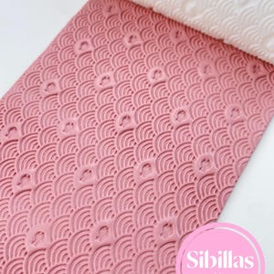 Pode incluir: Rolo de fondant rosa com um padrão de ondas repetidas. O padrão é gravado na superfície rosa. Uma seção branca enrolada do rolo mostra o mesmo padrão. O logotipo da Sibillas Cutters está no canto inferior direito.