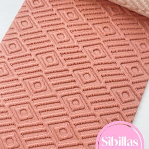 May include: A textured, coral-colored rolling pin with a repeating diamond pattern. The design is embossed into the surface, creating a raised relief. The rolling pin is partially rolled out, revealing a lighter, unrolled section. The text "Sibillas cutters" is visible.