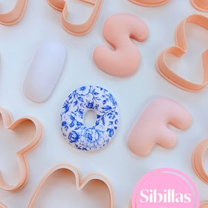 May include: A collection of peach-coloured plastic cookie cutters in various letter shapes, including a central letter O featuring a blue and white floral pattern. A pink circular logo with the white text Sibillas cutters is in the corner.