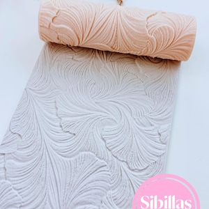Puede incluir: Un rodillo cilíndrico de color melocotón con un patrón en relieve complejo y arremolinado similar a hojas. Se coloca sobre una losa rectangular de color gris claro que muestra la misma textura orgánica, fluida y grabada. El texto Sibillas cutters es visible en un círculo rosa en la esquina inferior derecha.
