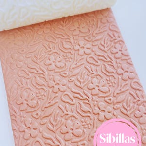 Può includere: Una lastra rettangolare per texture in argilla color pesca con un motivo in rilievo di vari fiori, foglie e forme organiche astratte. Un mattarello cilindrico bianco poggia sulla parte superiore. Un logo circolare rosa riporta Sibillas cutters.