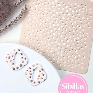 May include: A pale pink stencil with a pebble design and two square earrings with a white base and brown pebble design. The earrings have a square shape with a rounded inner opening. The image also includes a pink logo that says "Sibillas cutters".