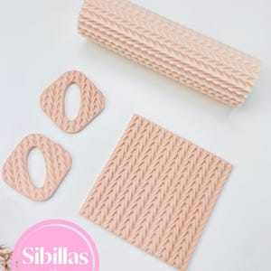 May include: A collection of light pink polymer clay tools for crafting. The set includes a textured rolling pin, two square-shaped cutters with oval cutouts, and a square textured sheet. The brand name "Sibillas cutters" is visible.