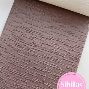 May include: A rectangular sheet of brown clay featuring a deeply embossed, horizontal, linear texture pattern. A white, textured roller is positioned at the top edge. A circular pink logo with the white text Sibillas cutters is in the bottom corner.