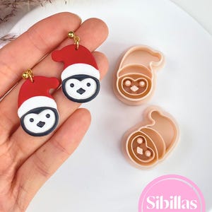 May include: Two penguin-shaped earrings featuring red and white Santa hats with gold-tone hardware, alongside two matching beige plastic cookie cutters. The image includes the text Sibillas cutters in a pink circle at the bottom right corner.