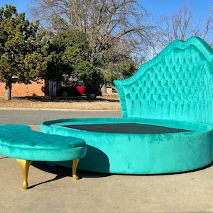 Puede incluir: Una cama redonda azul turquesa con un cabecero capitoné y un banco a juego azul turquesa con patas doradas. La cama está tapizada en terciopelo y tiene un colchón negro.