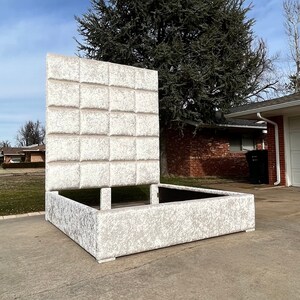 May include: A white upholstered bed frame with a tufted headboard. The headboard has a square pattern and is tall. The bed frame is sitting on a concrete driveway.