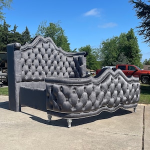 Puede incluir: Una cabecera capitoné gris con un diseño curvo y detalles plateados. La cabecera está unida a un piecero a juego con un diseño curvo. El marco de la cama está hecho de madera y tiene una tapicería de tela capitoné.