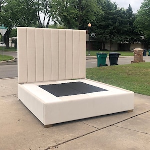 May include: A white upholstered bed frame with a tall headboard featuring vertical tufted panels. The bed frame has a square base and a black fabric insert.