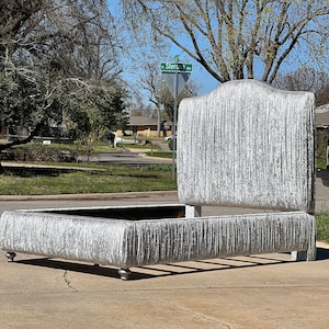 May include: A silver upholstered bed frame with a tall headboard and a tufted design. The bed frame is sitting on a concrete driveway.