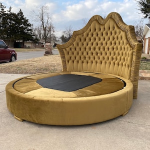 Puede incluir: Una lujosa cama redonda con cabecero acolchado y acabado de terciopelo dorado. La cama tiene un diseño único con una base circular y un centro negro. El cabecero tiene una parte superior decorativa festoneada.