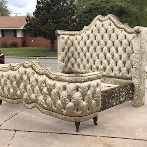 Puede incluir: Un armazón de cama tapizado y acolchado en color oro claro. El cabecero es alto y rectangular, con un piecero curvo. La tela tiene una textura similar al terciopelo y el marco se apoya en pequeñas patas oscuras.