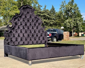 May include: A black velvet tufted headboard with a decorative, ornate design. The headboard is attached to a black velvet bed frame with clear acrylic legs.