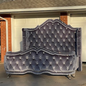 Puede incluir: Una cabecera capitoné de terciopelo gris con un patrón de diamante y detalles de cristal. La cabecera está unida a un marco de cama de terciopelo gris a juego con un diseño curvo y detalles de cristal.