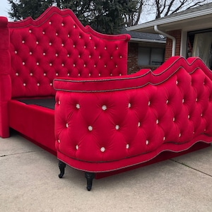 Puede incluir: Una cabecera tapizada de terciopelo rojo con un diseño curvo y botones de cristal. La cabecera está unida a un marco de cama de terciopelo rojo a juego con ribetes negros y patas negras.