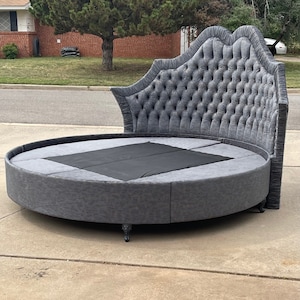 Puede incluir: Una cama redonda grande de color gris tapizada con un cabecero capitoné. El cabecero tiene un diseño festoneado y está cubierto de una tela suave y mullida.