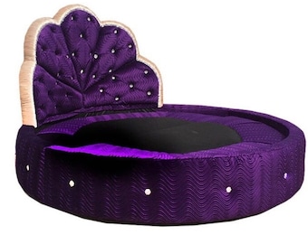 May include: A round, purple velvet bed with a large, ornate headboard. The headboard is covered in a purple velvet fabric with a quilted design and a gold trim.