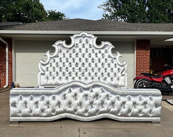 May include: A silver tufted headboard with a large, ornate design. The headboard is upholstered in a soft, plush fabric and features a curved shape.