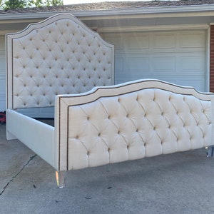 Puede incluir: Una cabecera capitoné blanca con la parte superior curvada y un ribete decorativo. La cabecera está unida a un marco de cama blanco con patas de acrílico transparente.