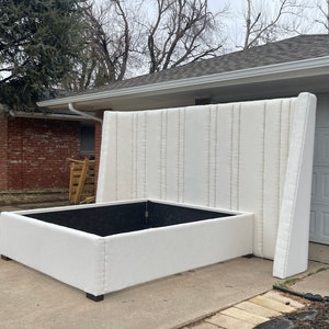 May include: A white upholstered bed frame with a tall headboard featuring vertical tufted panels. The bed frame is sitting on a concrete driveway.